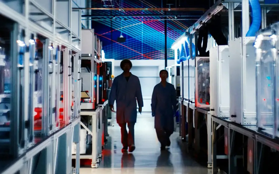  Two people walk in an aisle in an industrial-looking lab.