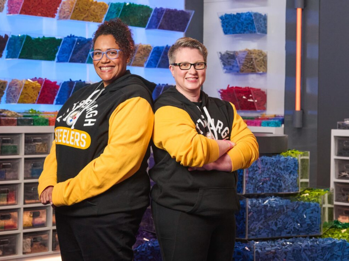  Two women stand back to back in front of bins filled with LEGO bricks.