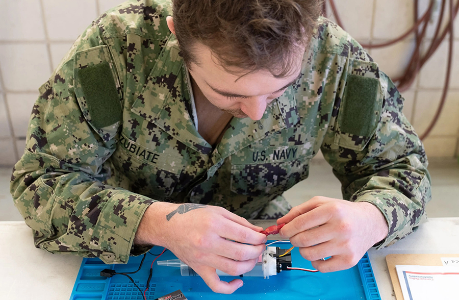  A man wearing camoflage works at a desk, wiring a small robotic device.