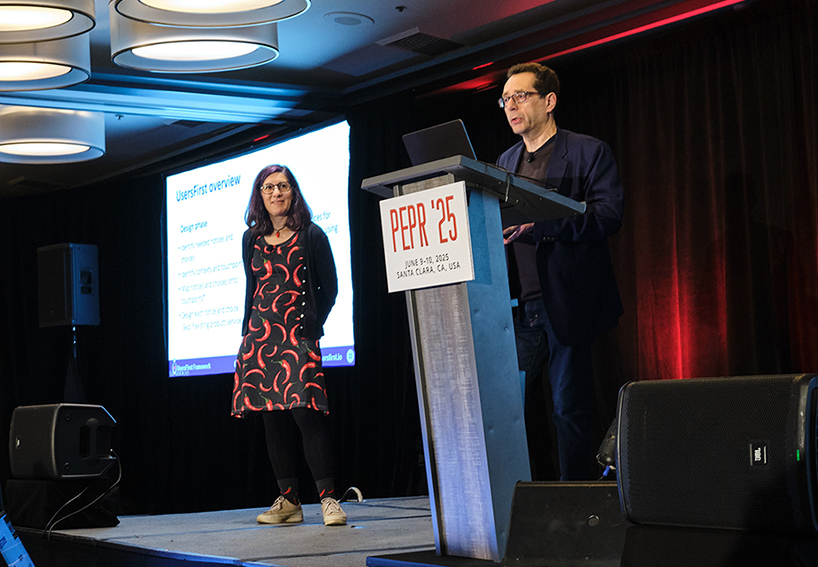  A man stands behind a podium, speaking, while a woman in a dress covered in red chili peppers watches.